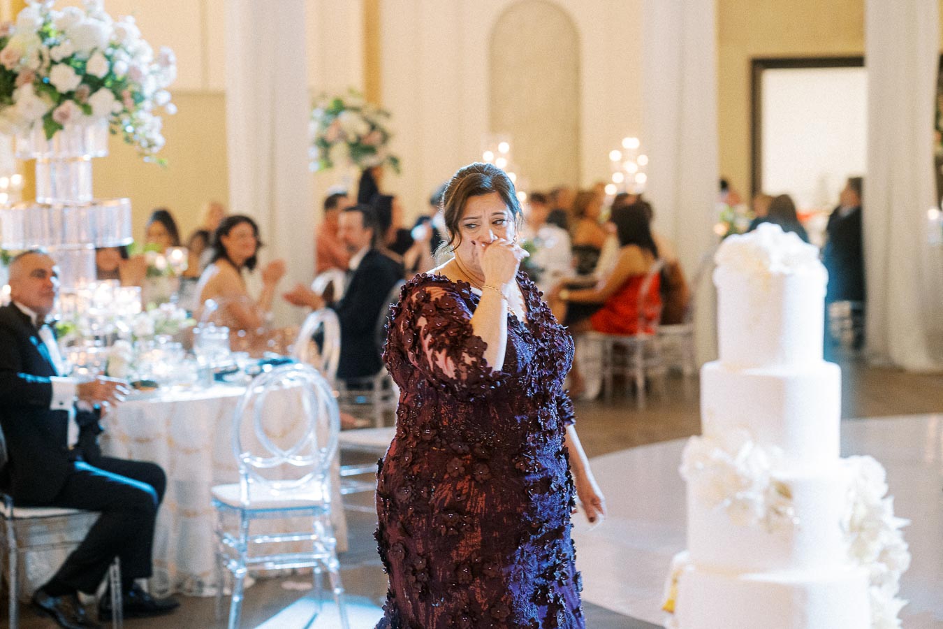 A woman in a purple dress emotionally reacts at a wedding reception, surrounded by elegantly decorated tables and a tiered