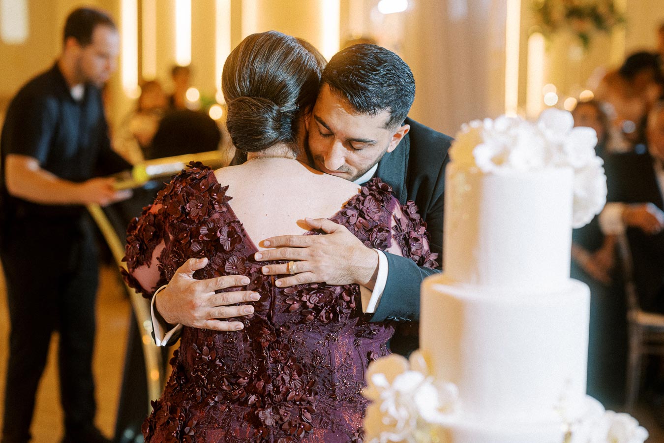 A heartfelt embrace between a man in a suit and a woman in a floral burgundy dress during a wedding celebration, with a