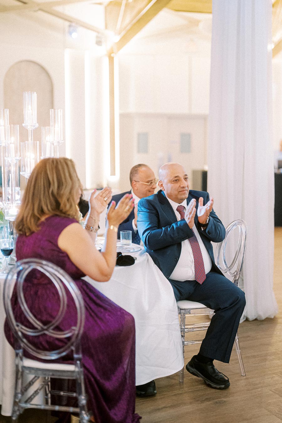 Elegant wedding reception with guests in formal attire clapping at a beautifully set round table adorned with candle