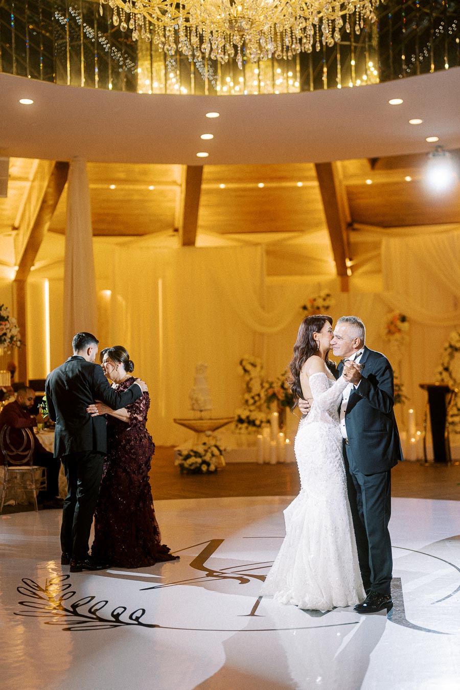 A beautifully decorated wedding reception hall with two couples dancing under an elegant chandelier. The bride and groom,