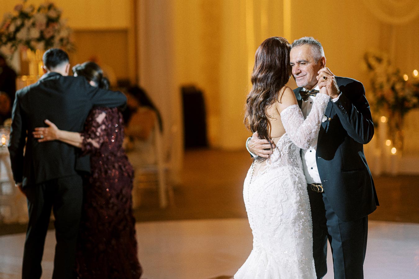 A bride in a white lace gown dances with an older man at a wedding reception, with another couple embracing in the