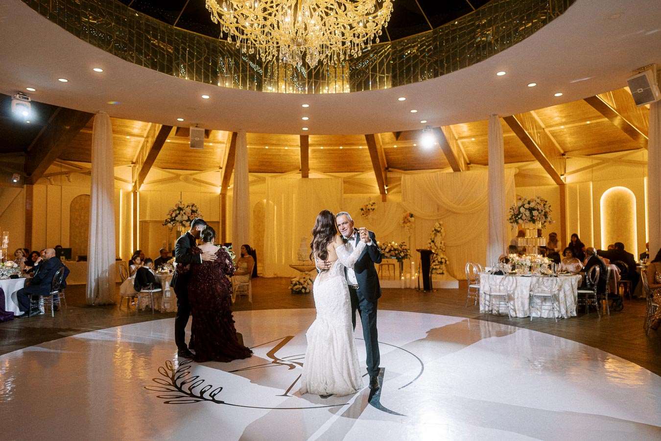 A bride and groom share their first dance under a stunning chandelier in an elegantly decorated ballroom, surrounded by