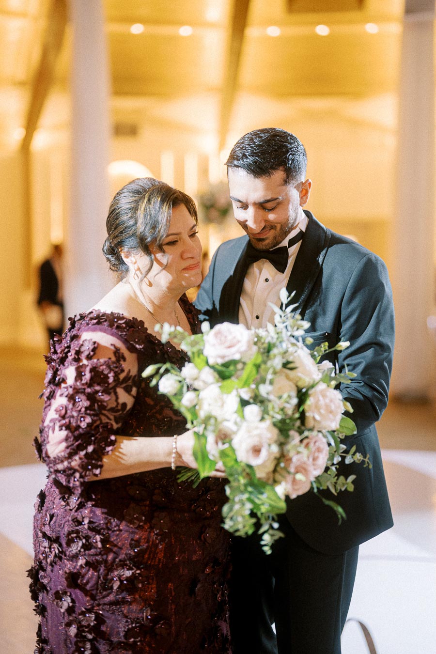 A joyous wedding moment captured as a man in a tuxedo embraces a woman in an elegant burgundy dress, holding a lush bouquet