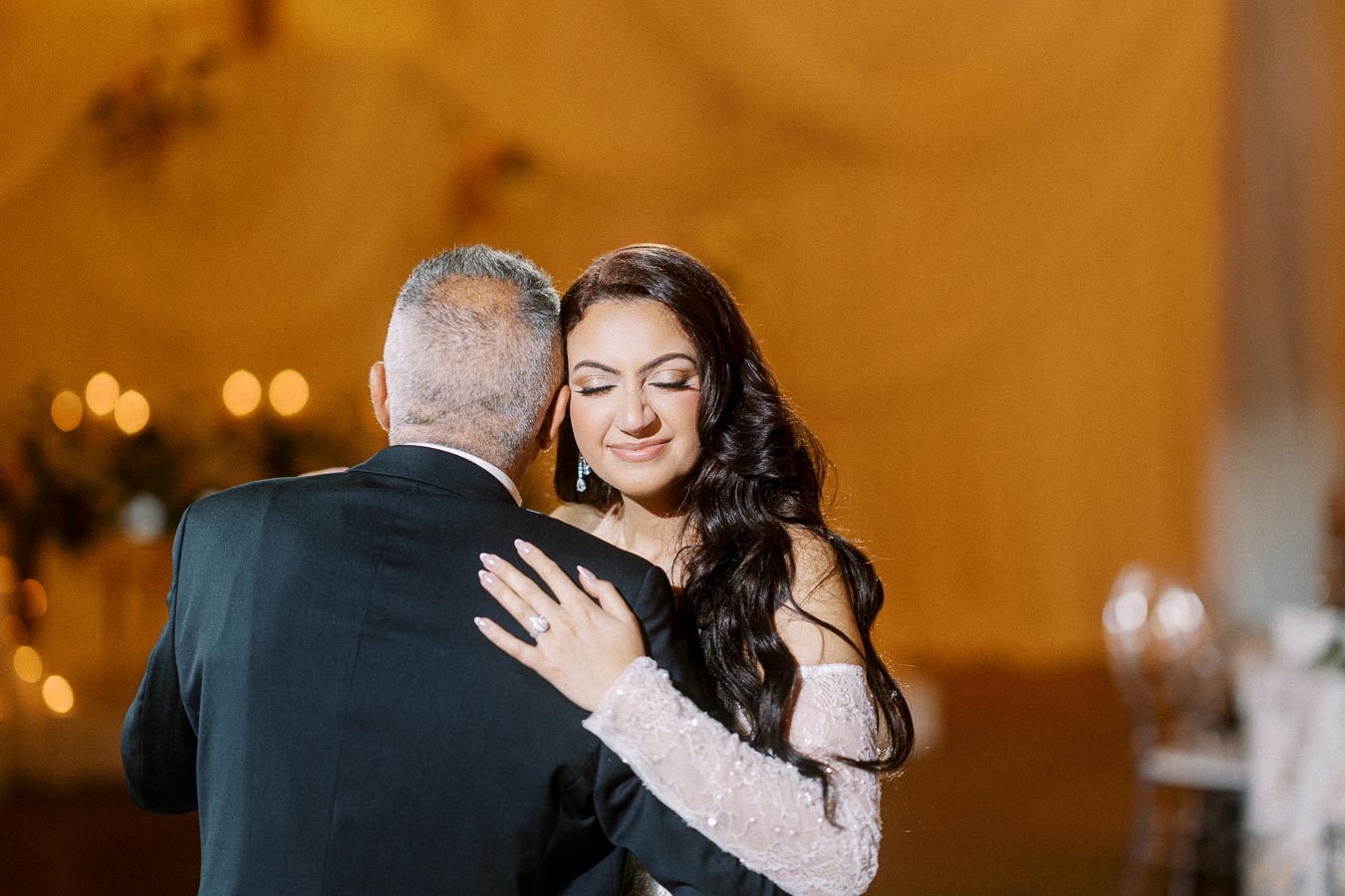 A bride in a lace gown embracing an older man in a suit, likely during a wedding dance, with a soft, warmly lit background.