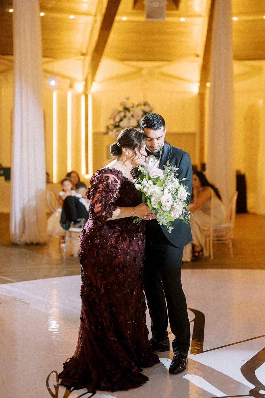 A groom in a black tuxedo shares a heartfelt dance with a woman in a burgundy dress, holding a bouquet of white and pink