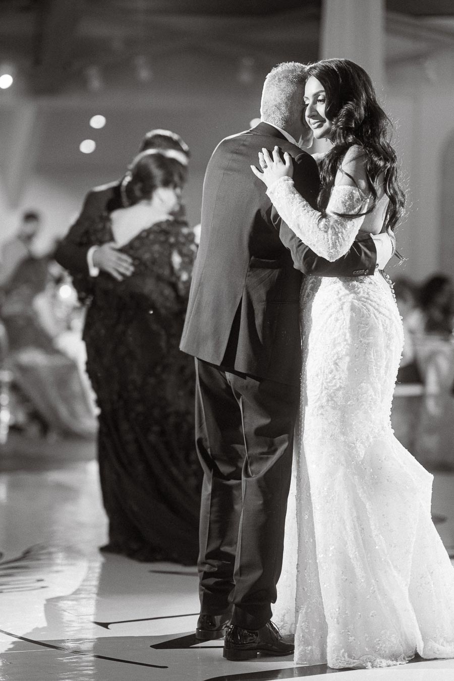 A bride in a white lace gown gracefully dancing with her father at a wedding reception, captured in a warm, intimate
