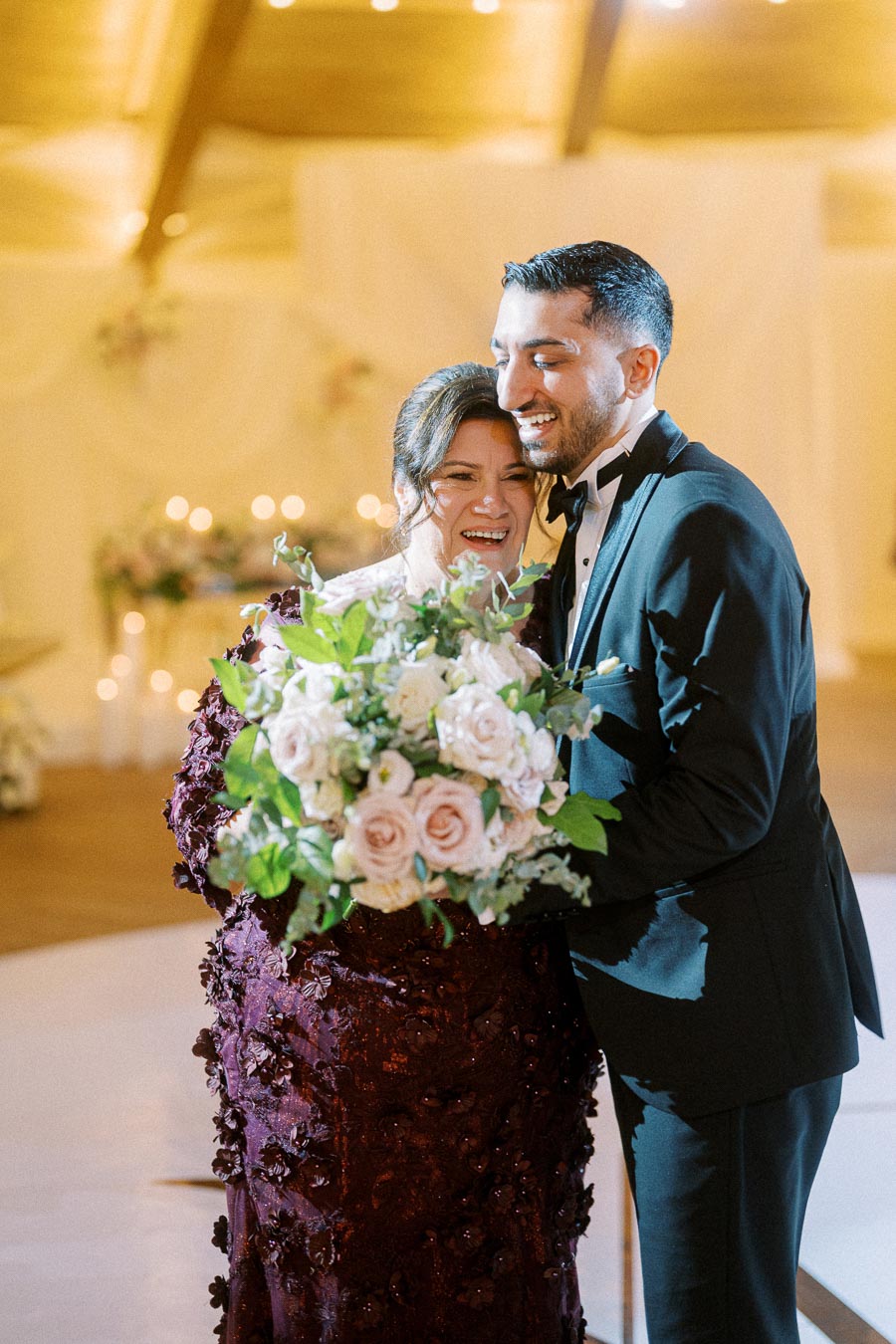 A joyful couple embracing at their wedding reception, with the bride holding a bouquet of pink and white roses and the groom