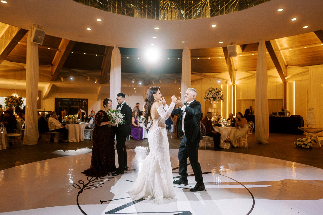 A bride in a white gown dances with her father on a spacious, elegantly decorated wedding reception dance floor, surrounded