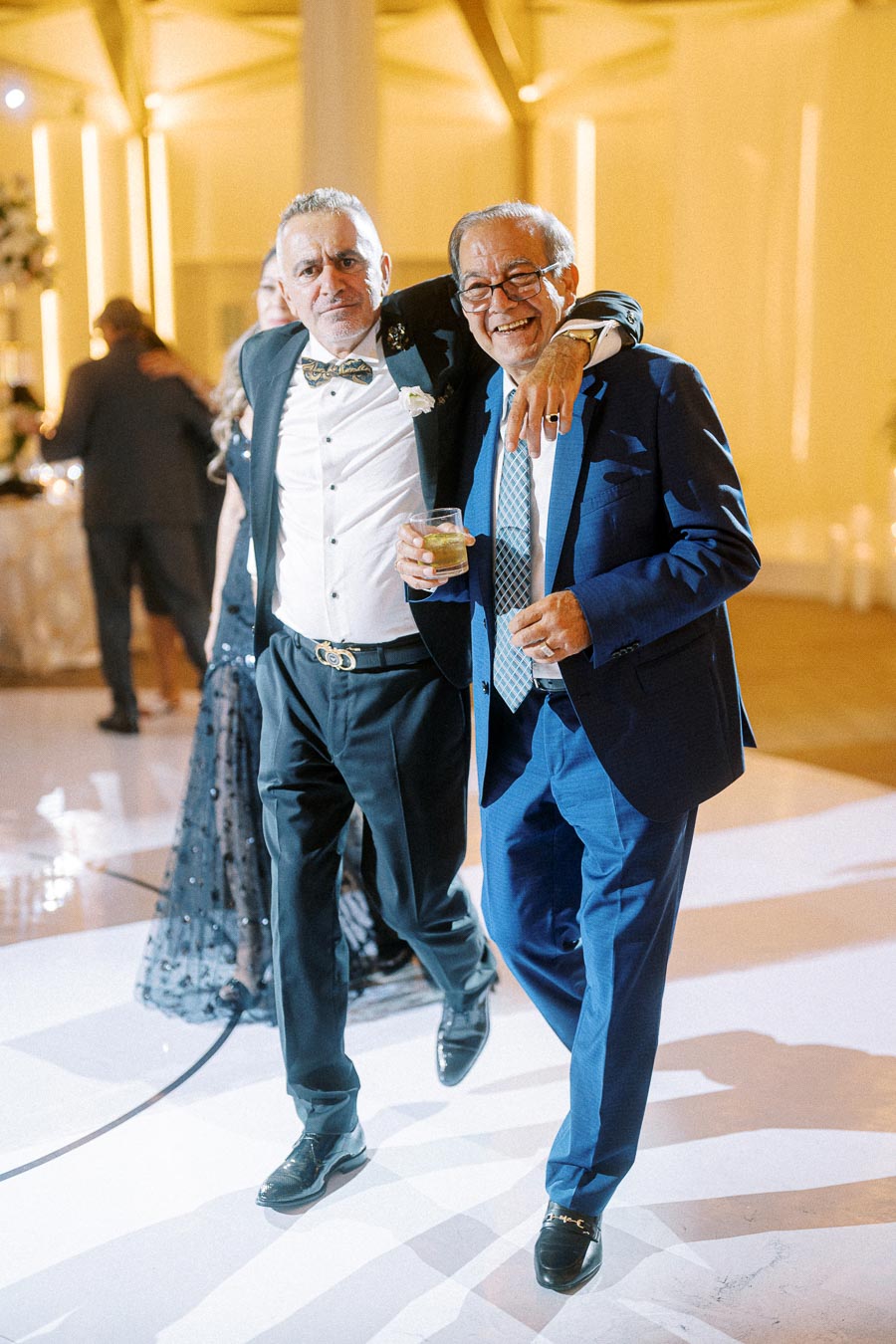 Two men in suits enjoying a celebration at an elegant event, one with an arm around the other, both holding drinks and