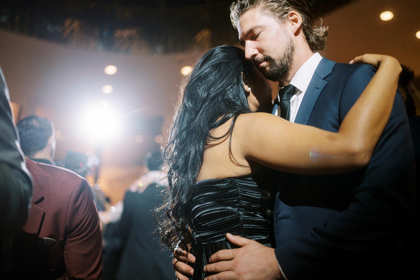 A couple in formal attire sharing a romantic embrace during a dance at a wedding reception, with soft lighting and guests