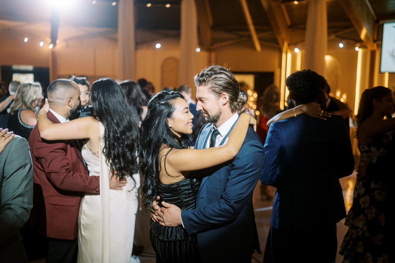 A couple dancing closely at a lively wedding reception, surrounded by other joyful guests on the dance floor, under elegant