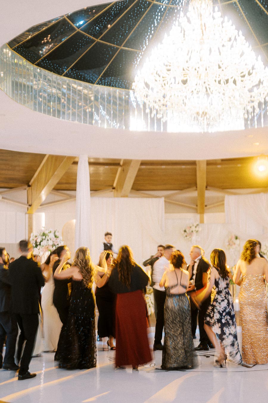 Wedding reception with elegantly dressed guests dancing under a grand chandelier in a spacious venue.
