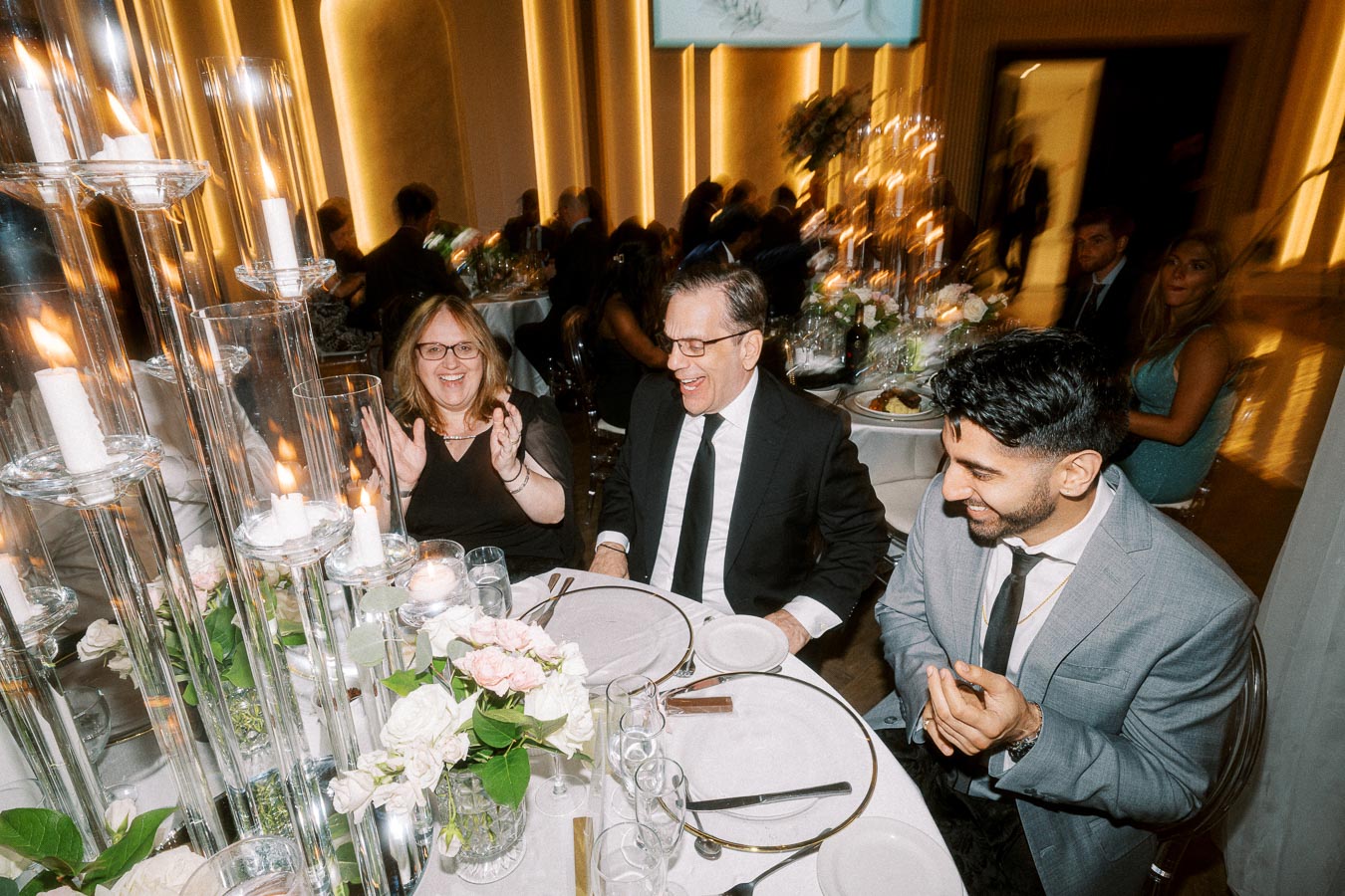 Three people in formal attire are smiling and engaged in conversation at an elegantly decorated dinner table with floral