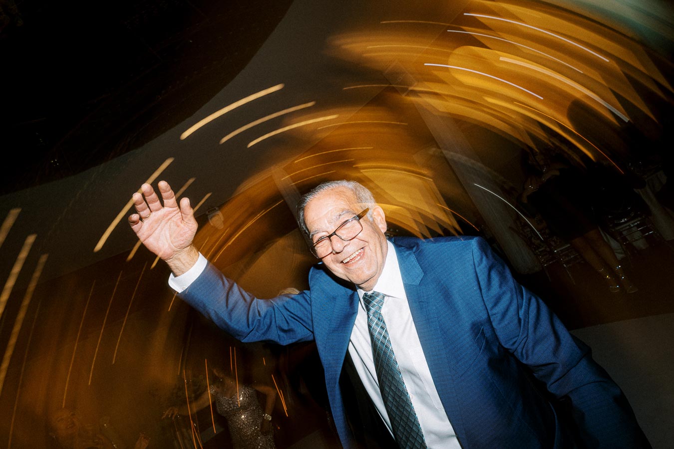 Elderly man in a blue suit and glasses joyfully dancing at an event, with a blurred background of lights creating a dynamic,