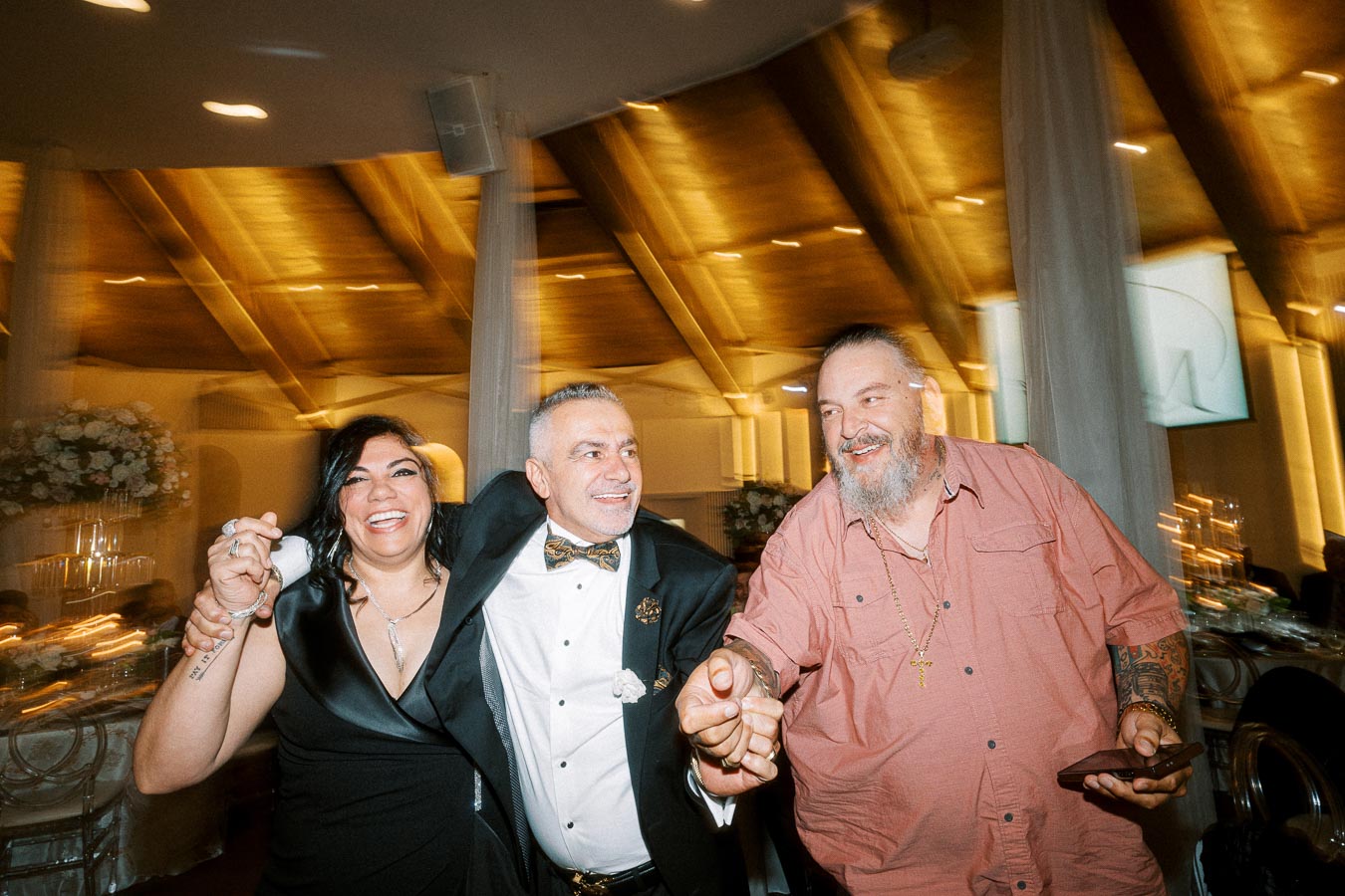 Three joyful people celebrating at a wedding reception in a warmly lit venue.