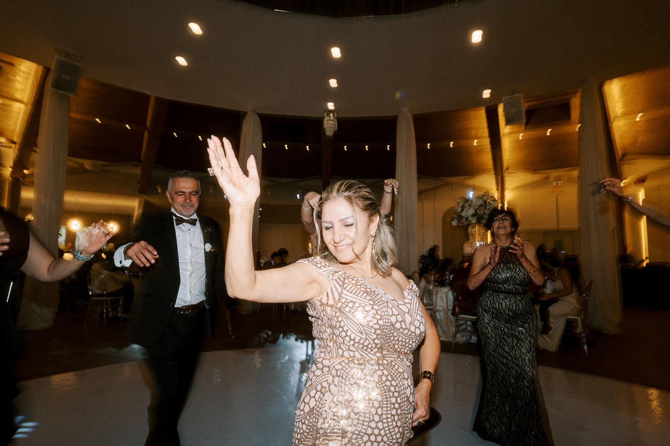 Group of elegantly dressed people dancing joyfully at a formal event with ambient lighting and decorated venue.