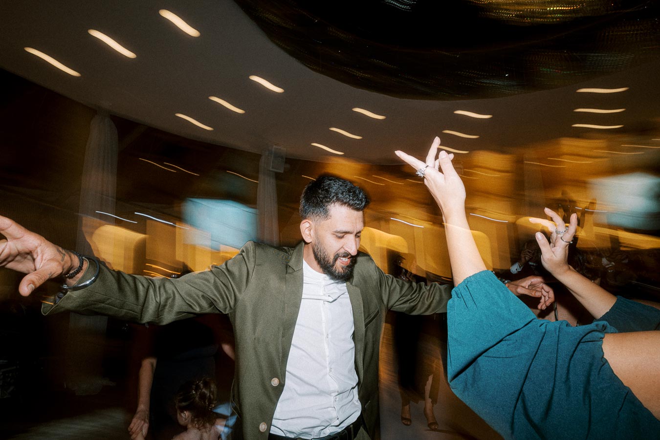 A man in a green suit energetically dancing at a party with blurred motion lights in the background.