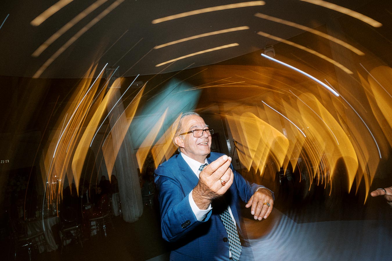 Elderly man in a blue suit dancing joyfully at a party, surrounded by dynamic, swirling light trails creating an energetic