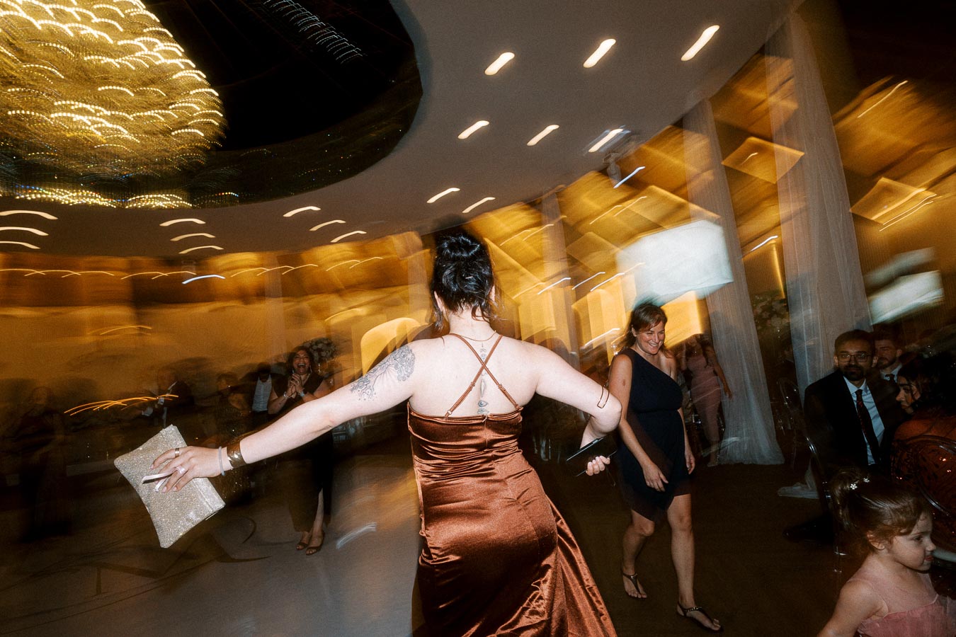 A woman in a flowing brown dress joyfully dances at a lively indoor celebration, carrying a glittery clutch purse. The room