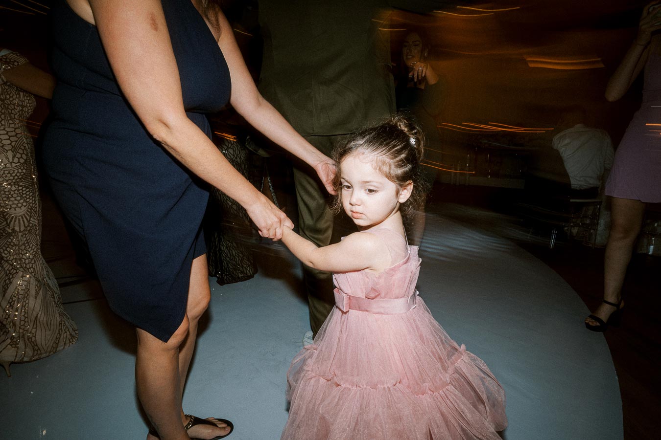 A young girl in a pink dress holds hands with an adult in a navy dress at a lively indoor event.