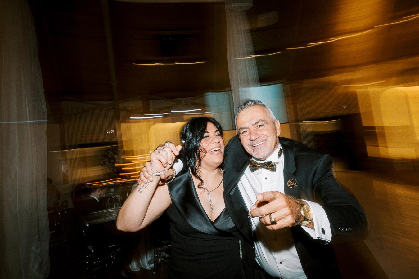 A joyful couple dancing at an evening event, dressed in formal black attire, showing happy expressions with motion blur in