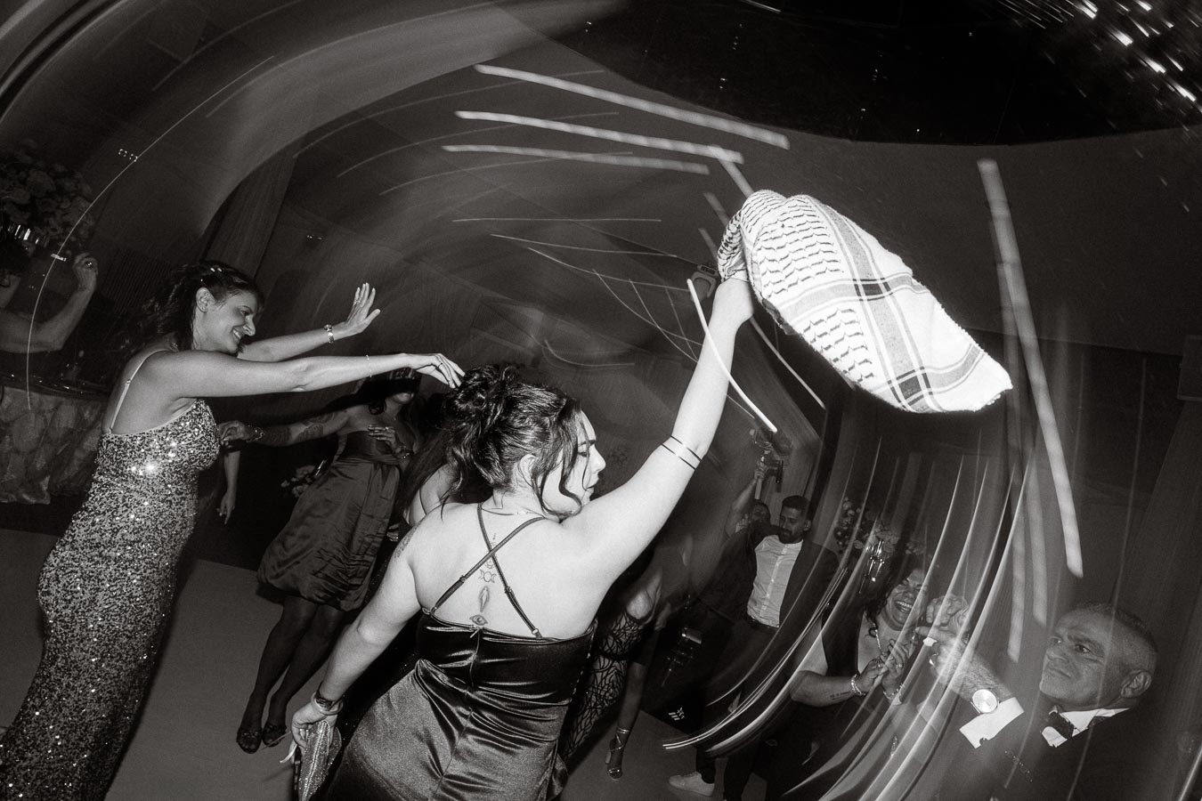 Black and white image capturing a lively dance floor scene at a celebration, featuring elegantly dressed women waving