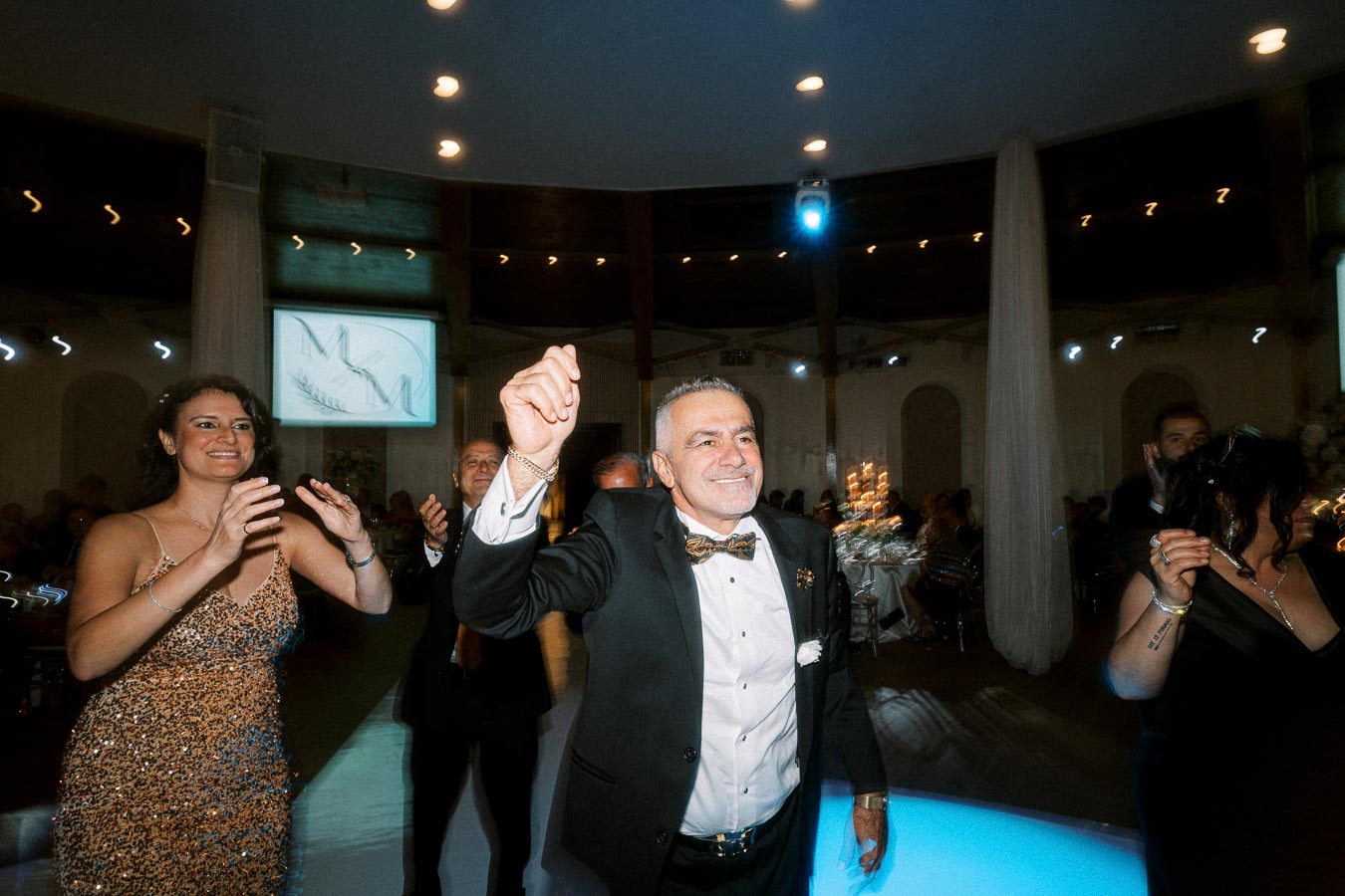 A joyous wedding reception scene with guests dancing in an elegant venue; a man in a tuxedo smiling while leading a lively