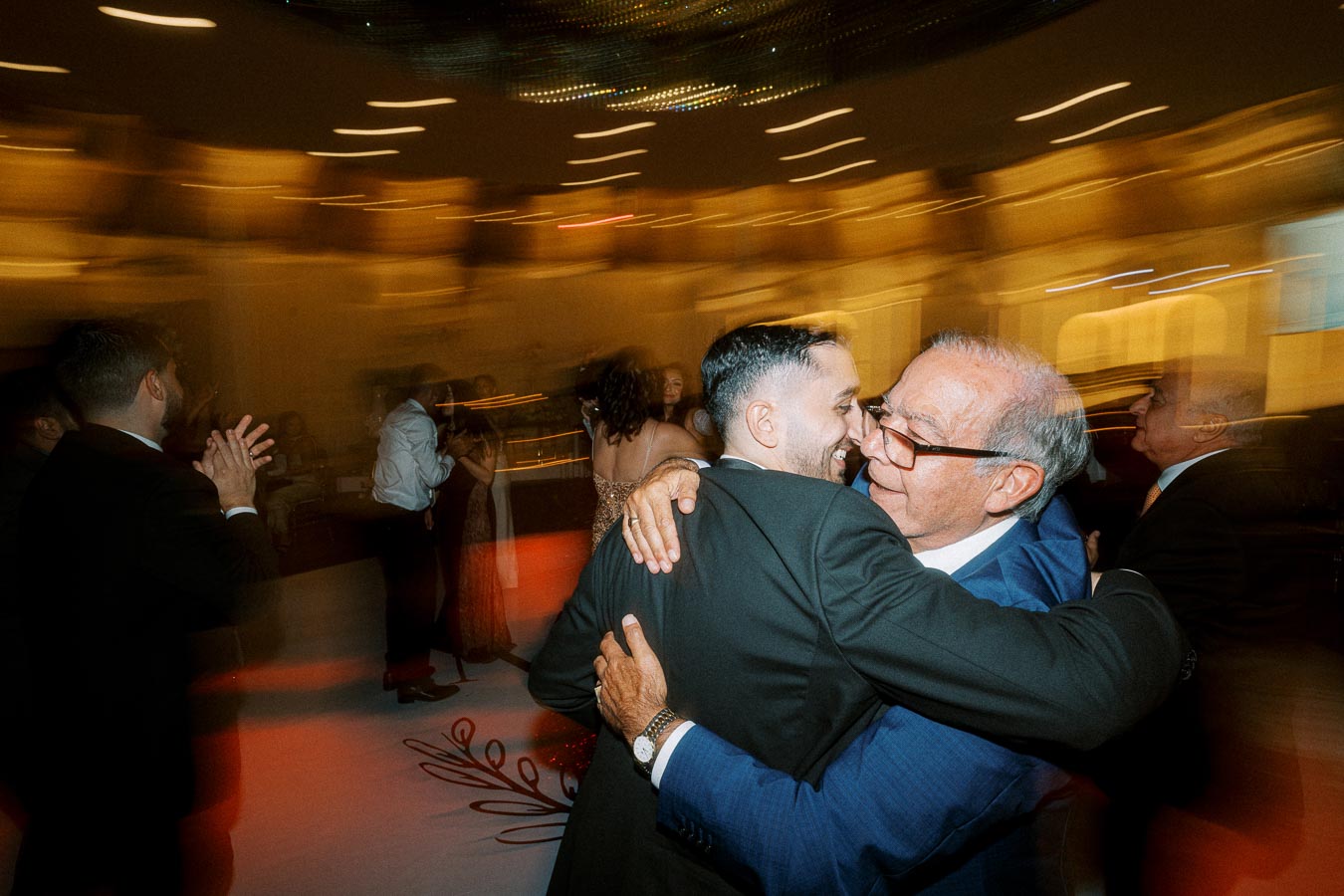 A joyful moment at a wedding reception with two men embracing warmly on the dance floor, surrounded by guests in a softly