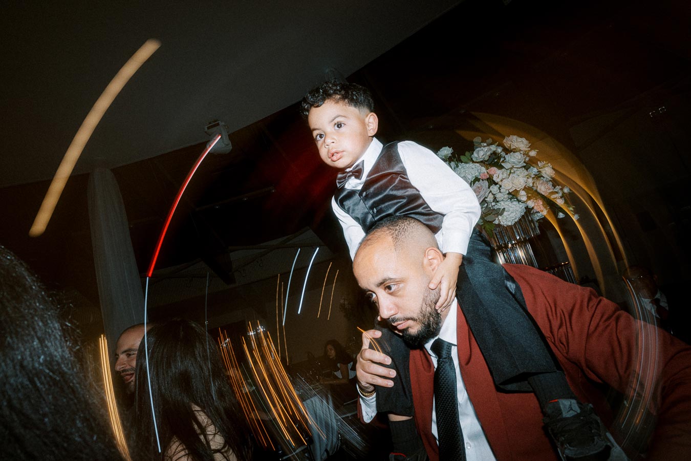 A man in a red jacket carries a child in a white dress shirt and black vest on his shoulders during a lively indoor event,