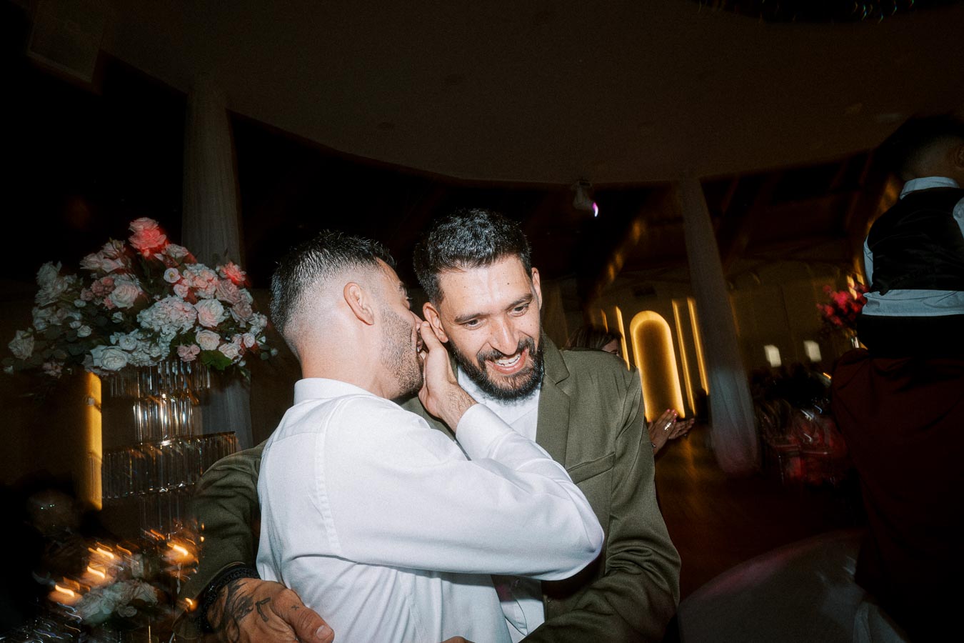A candid moment of two friends embracing and sharing a secret at a wedding reception, with floral arrangements and warm