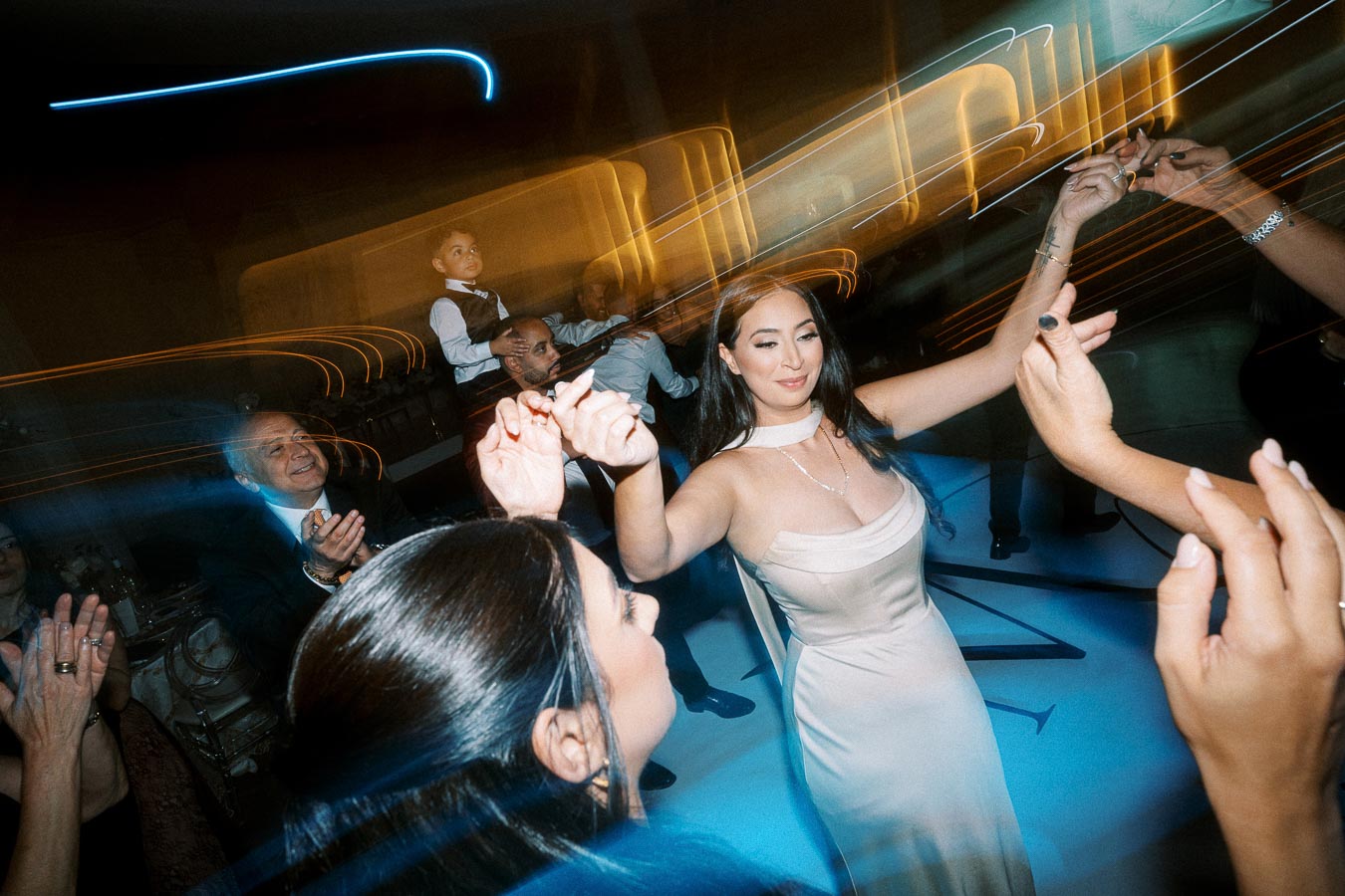A woman in an elegant beige dress dances joyfully with guests at a lively evening celebration, surrounded by vibrant light
