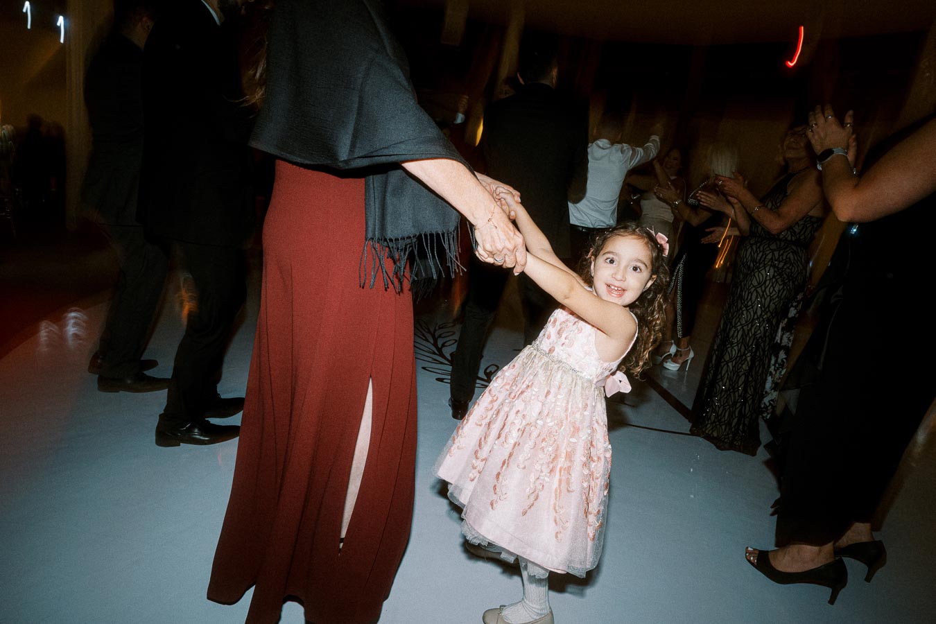 A young girl in a pink dress joyfully dances with an adult at a lively, dimly-lit celebration or event, surrounded by people