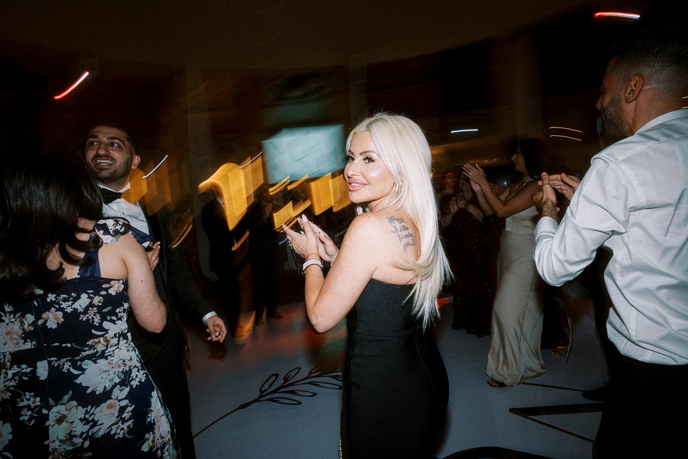 A woman with long blonde hair and a visible tattoo on her shoulder smiles at the camera while dancing at a lively party,