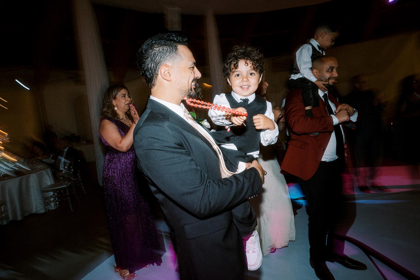 A joyful celebration scene featuring a well-dressed man holding a smiling child with curly hair, as another adult carries a