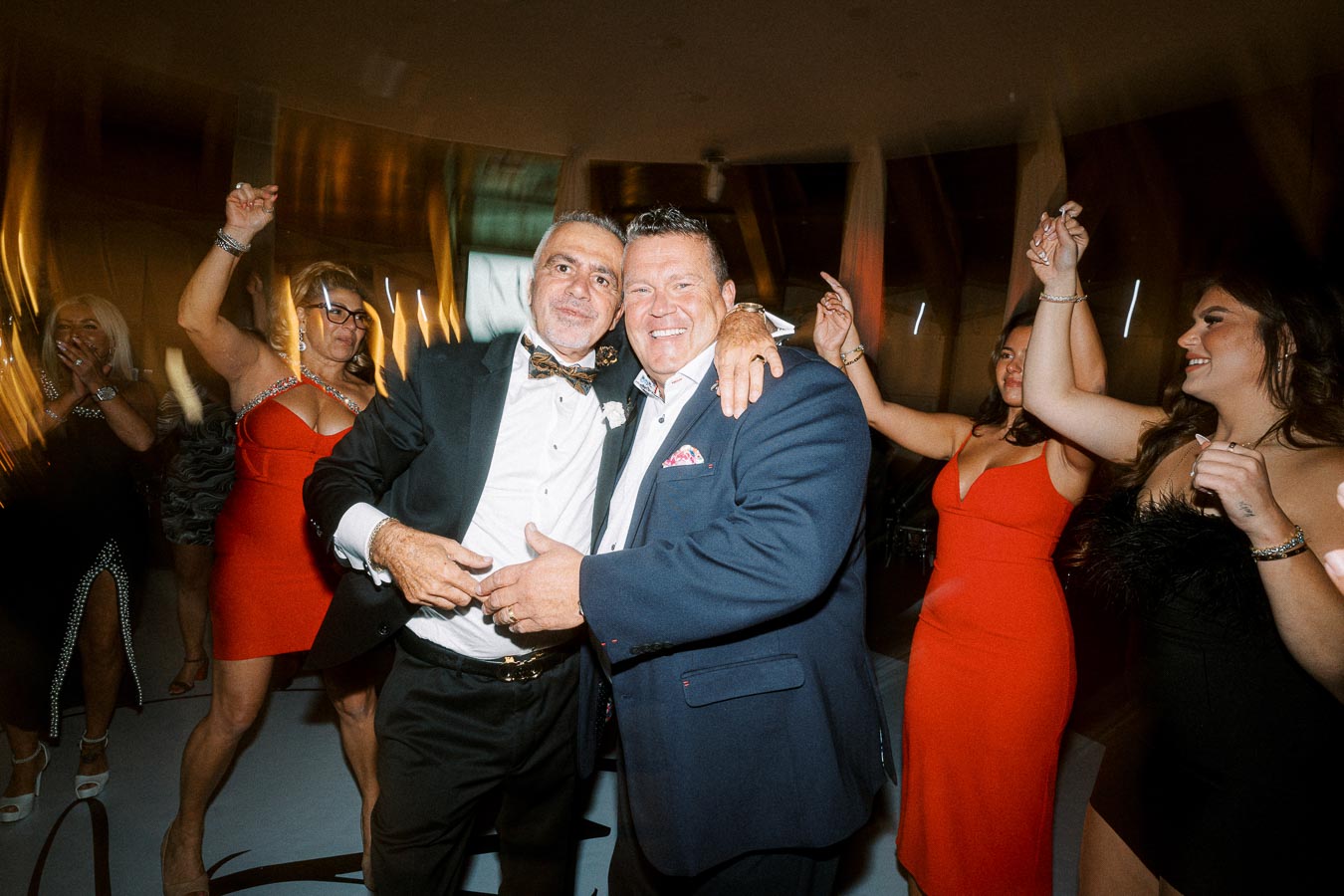 A lively dance floor scene at a formal event, featuring two well-dressed men smiling and posing together, surrounded by