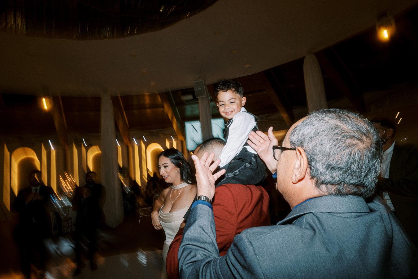 A joyful scene at a celebration with a smiling young boy on a man's shoulders, surrounded by happy adults in a warmly lit