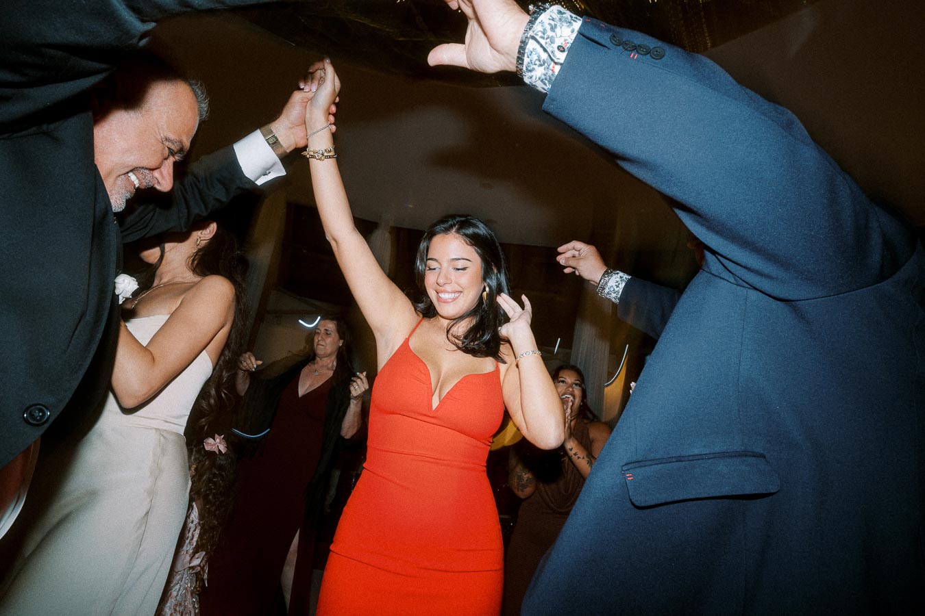 A joyful woman in a red dress dancing at a lively indoor celebration, surrounded by smiling and applauding guests.