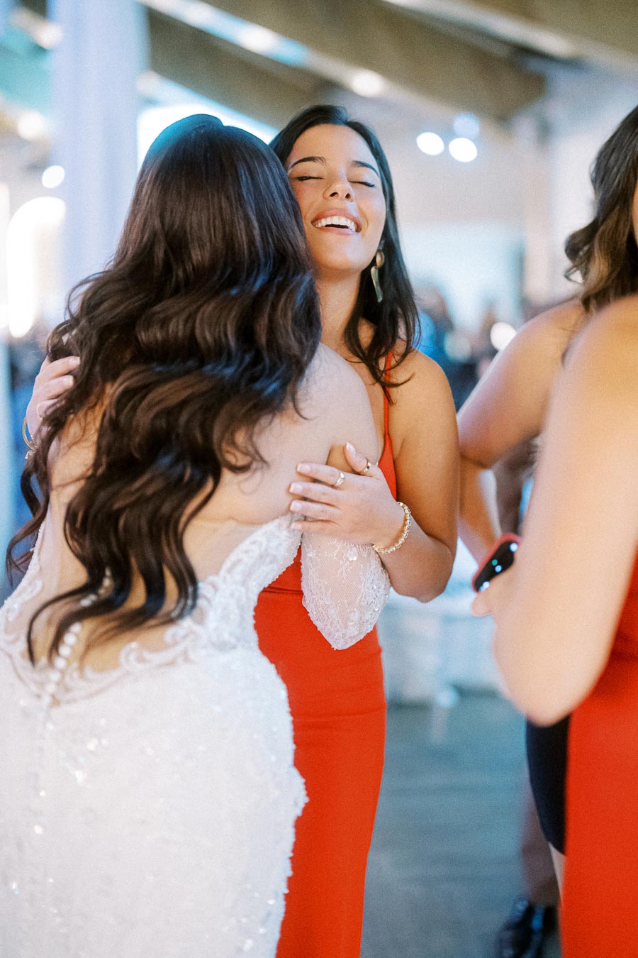 Two women embracing at an event, one in a white lace dress and the other in a red gown, conveying joyful and celebratory