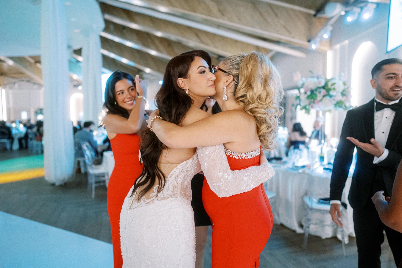 A bride in a white gown embraces a guest in a red dress at a wedding reception, with guests around them in formal attire.