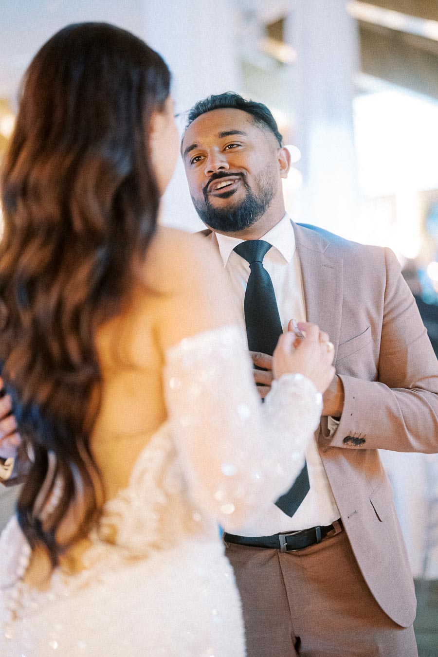 A groom in a tan suit and black tie smiles while dancing with his bride in a white wedding dress during their wedding