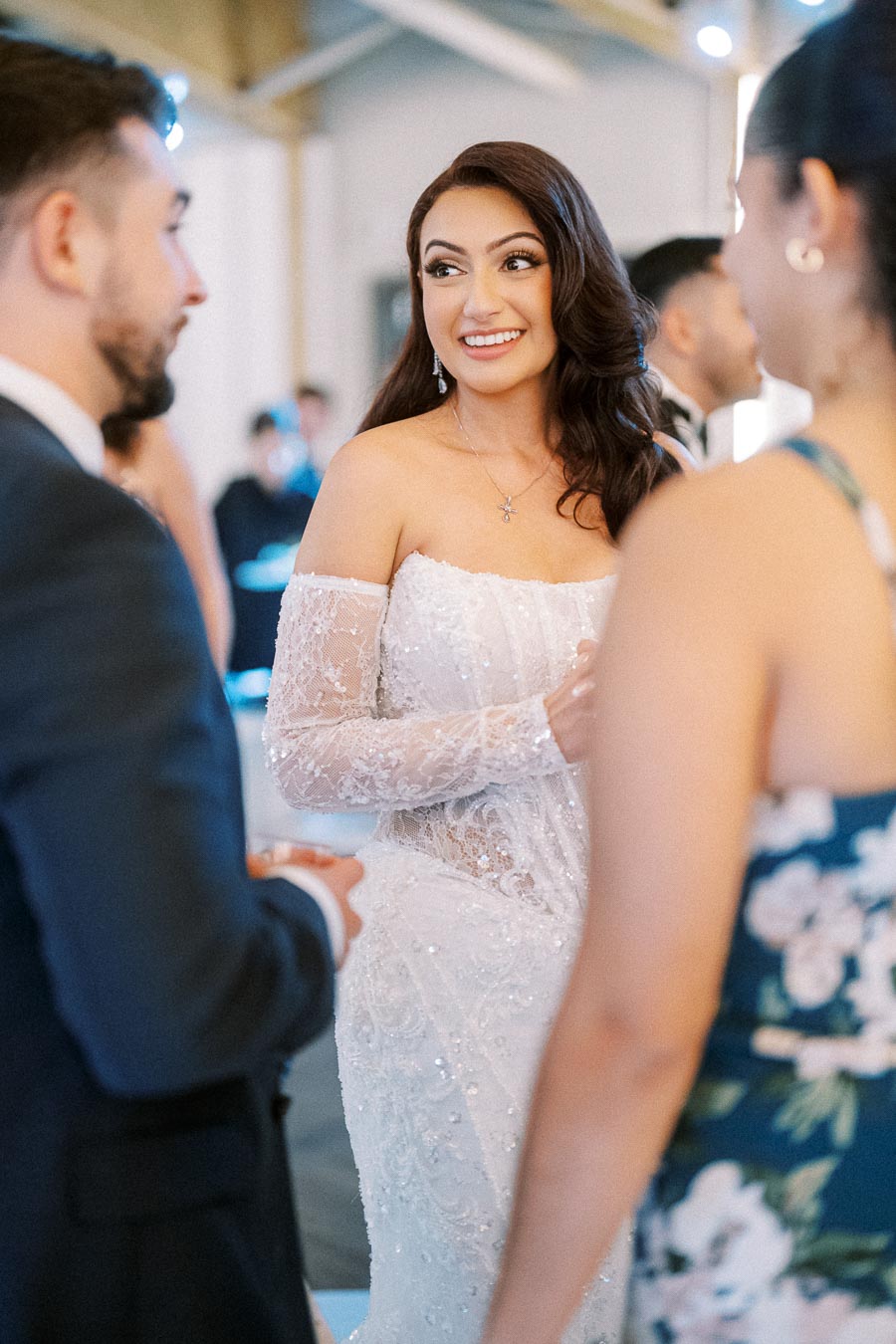 A bride in an elegant white lace gown smiling while talking to guests at a wedding reception.