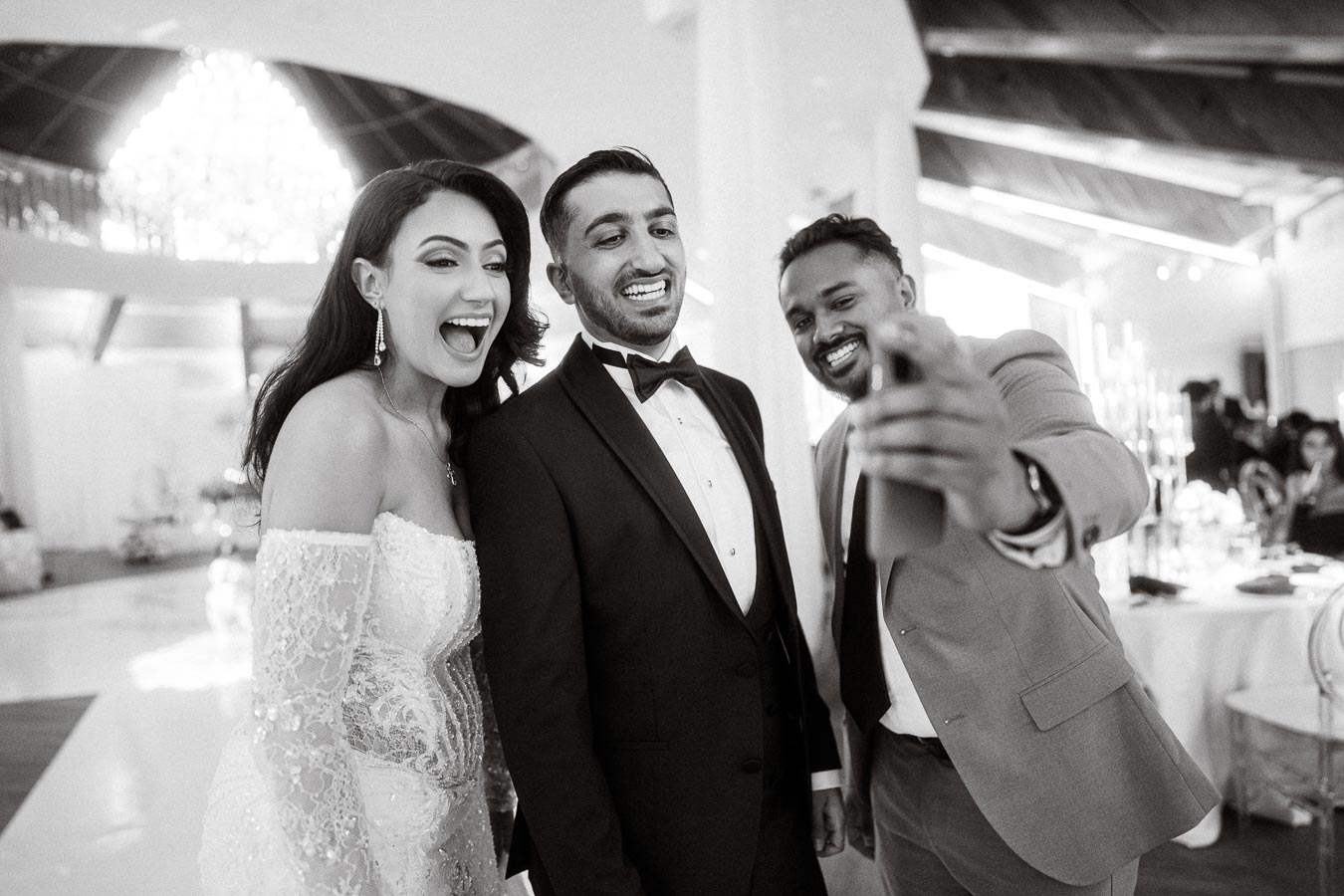 A happy bride and groom in elegant wedding attire take a joyful selfie with a friend at a stylish indoor wedding reception.