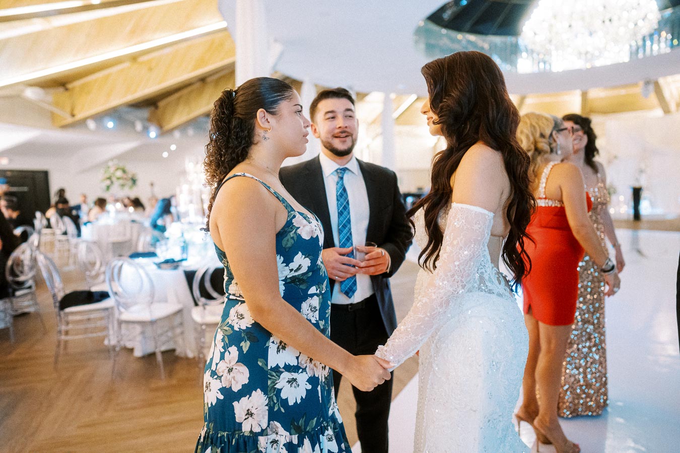 Elegant indoor wedding reception scene with a bride in a lace gown, holding hands with a guest in a floral dress, and a man