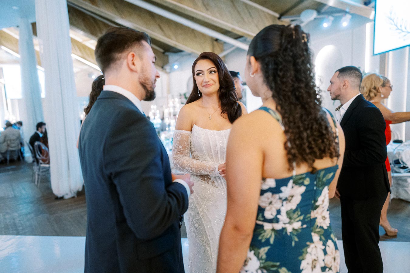 A bride in an elegant white wedding dress with lace sleeves is happily conversing with guests at a wedding reception. The