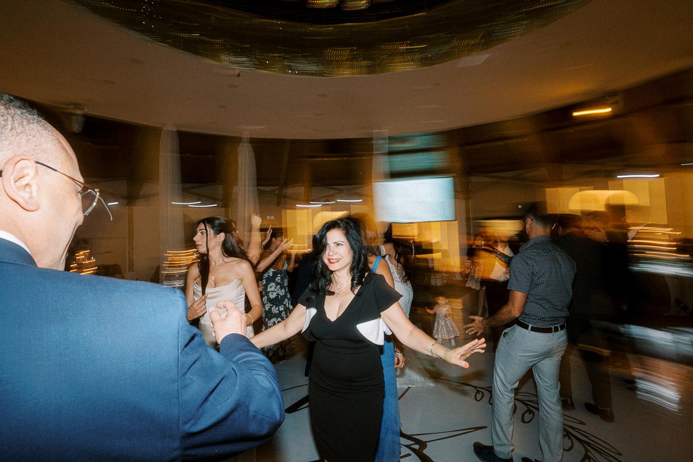 A lively party scene with guests dancing energetically under dim lighting, featuring a woman in a black dress in the