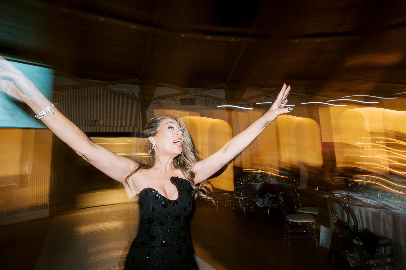 A woman joyfully dancing in a dimly lit venue, wearing an elegant black dress, with motion blur creating a dynamic and
