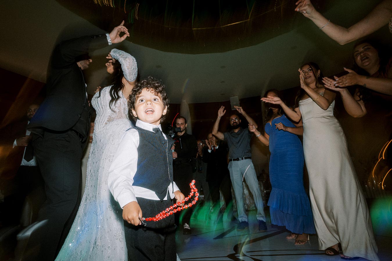 A joyful wedding celebration with a child in formal attire holding a red beaded necklace, surrounded by guests dancing and