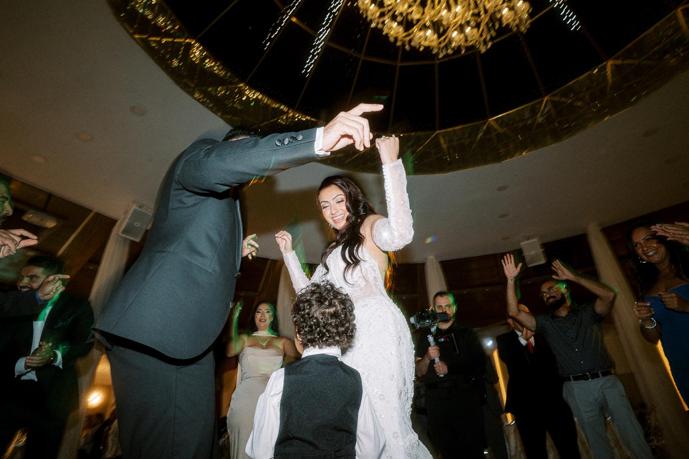 A joyful wedding celebration under a grand chandelier, featuring a bride in a white gown dancing with guests in formal