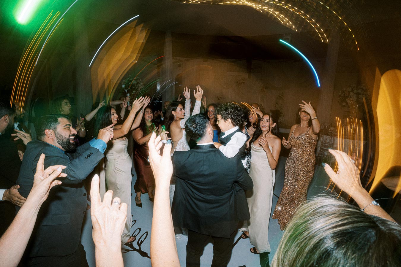 A lively wedding reception with guests dancing energetically, a man holding a child, surrounded by elegantly dressed women
