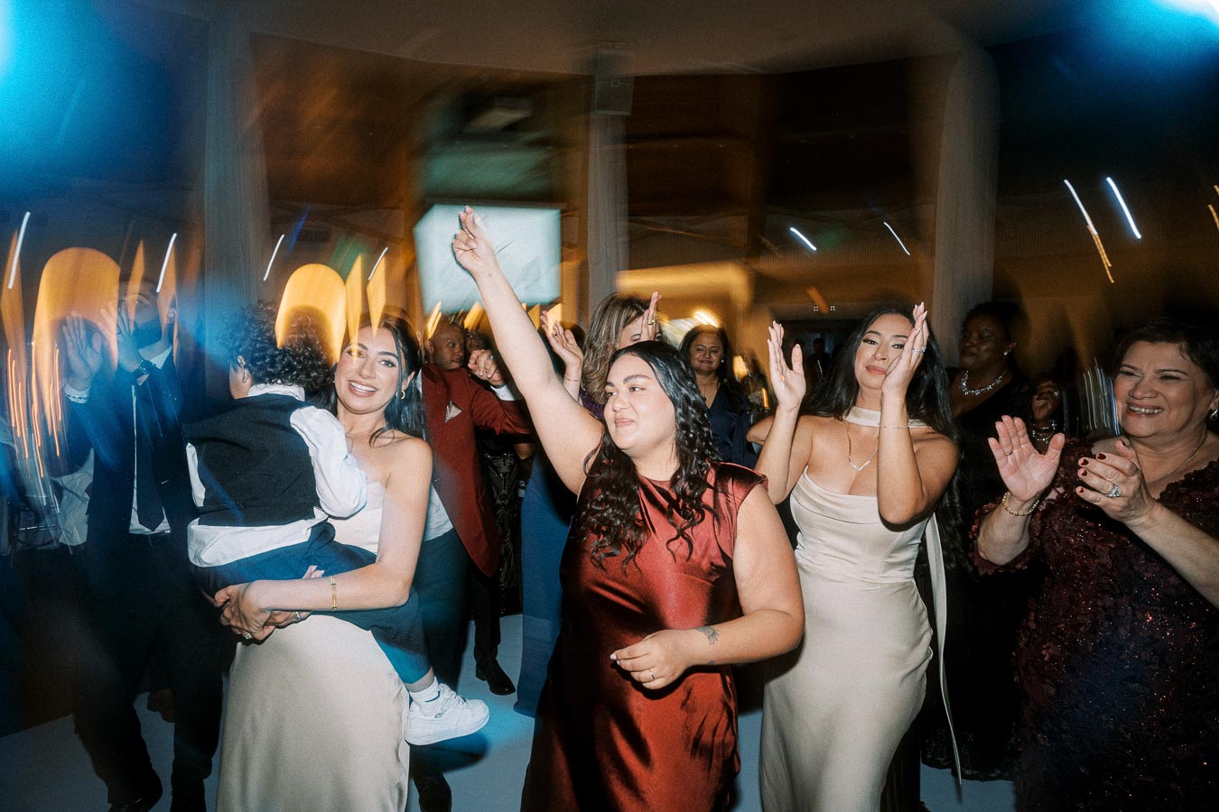A joyful wedding celebration with guests dancing and clapping, featuring a woman holding a child, others in elegant dresses,
